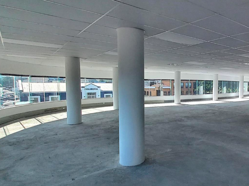 An expansive, open-plan office space featuring a suspended ceiling, structural columns, and large wrap-around windows offering natural light and street views.
