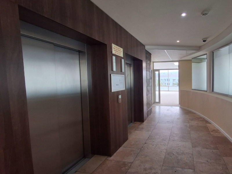 A modern office building hallway featuring dual elevators, stone flooring, and recessed lighting, leading to a glass-enclosed exit area.