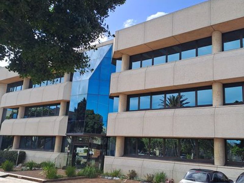 A multi-story office building featuring a modern glass facade, concrete paneling, tinted windows, and a paved entrance area with landscaped garden beds.