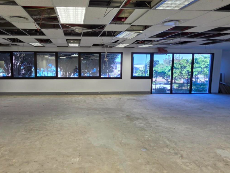 Spacious open-plan office area featuring large windows, concrete flooring, and a suspended ceiling grid currently undergoing renovation with exposed wiring and missing tiles.
