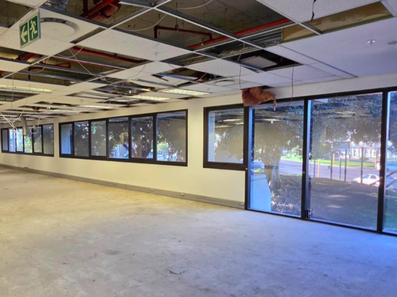 An open-plan office space featuring large windows, exposed ceiling infrastructure, and missing ceiling tiles, currently in a shell condition ready for renovation.