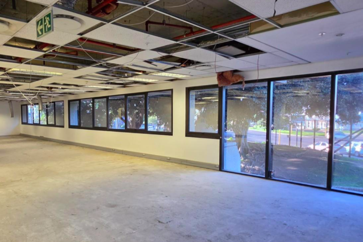 An open-plan office space featuring large windows, exposed ceiling infrastructure, and missing ceiling tiles, currently in a shell condition ready for renovation.