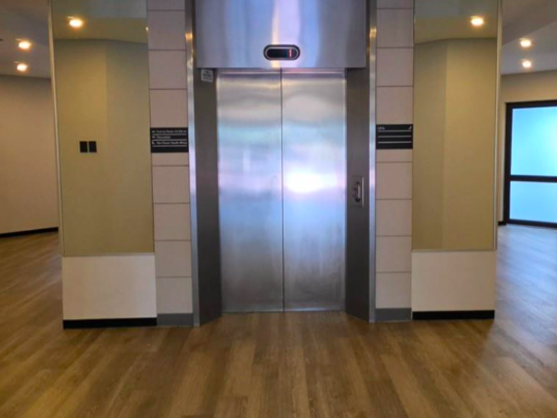 A clean and modern office building elevator lobby featuring stainless steel elevator doors, wood-look flooring, and mirrored wall accents.