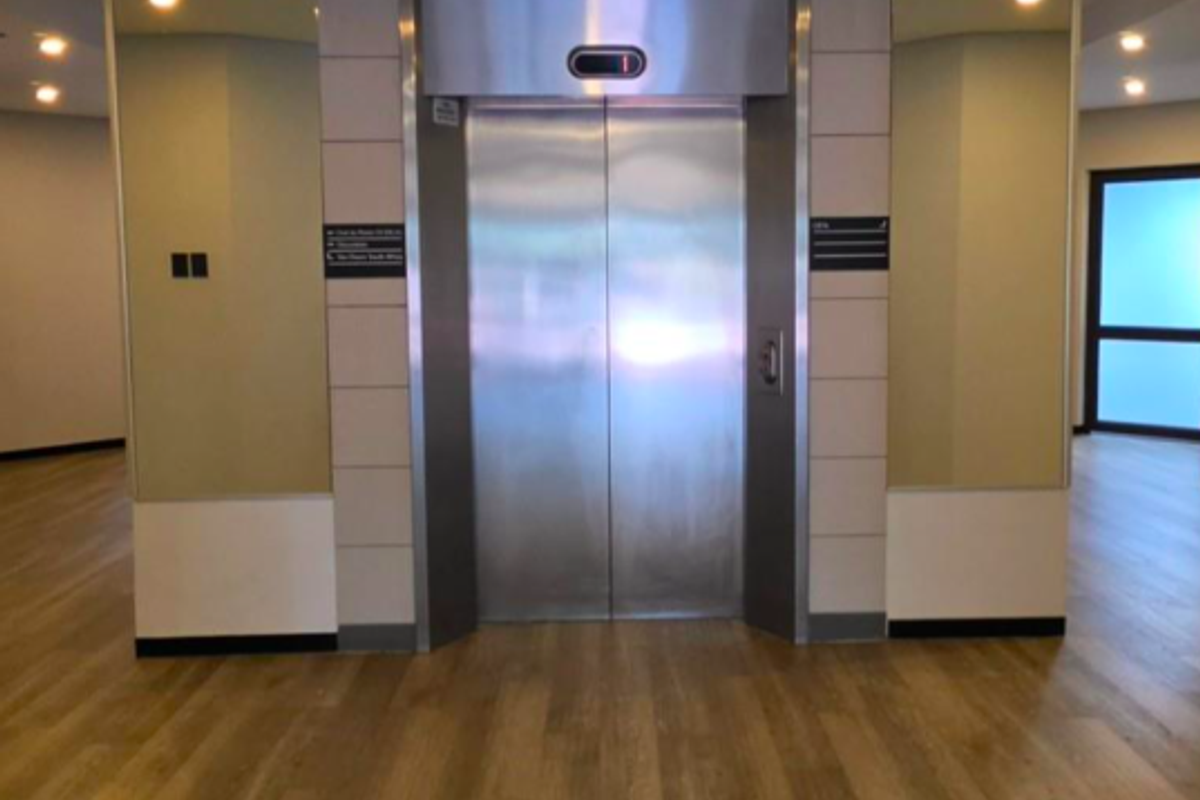 A clean and modern office building elevator lobby featuring stainless steel elevator doors, wood-look flooring, and mirrored wall accents.