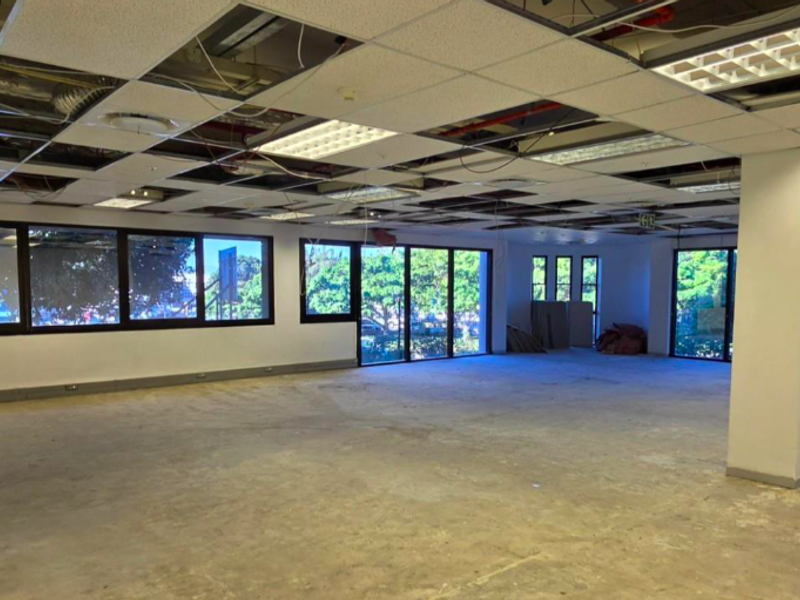 An expansive, open-plan office space featuring large windows, a structural column, and a partially dismantled suspended ceiling requiring renovation.