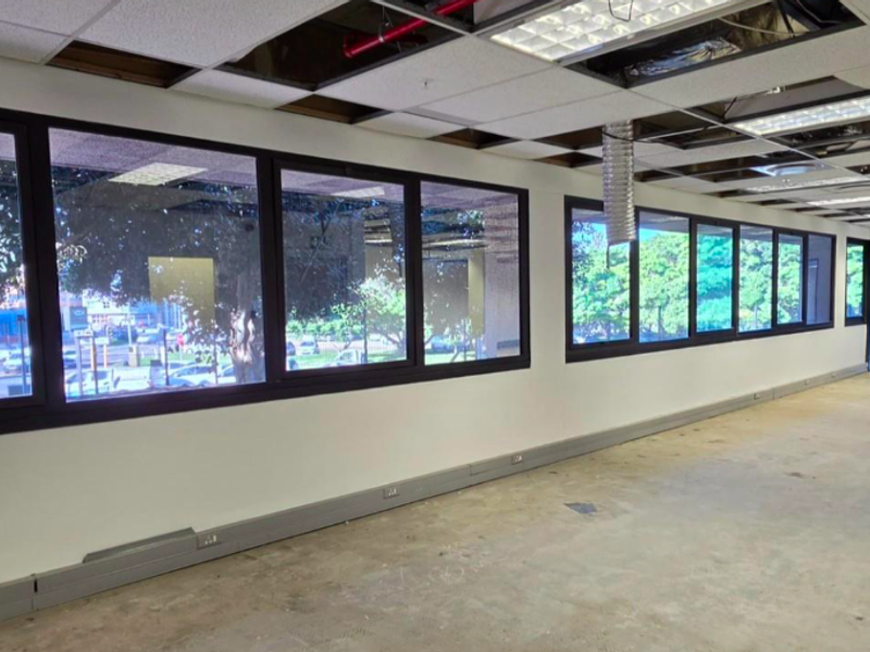 An expansive, open-plan office space currently undergoing renovation, featuring large windows, perimeter trunking, and an exposed ceiling grid with missing tiles.