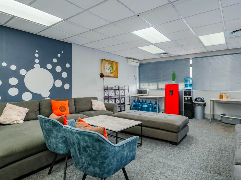 A functional office break room featuring a suspended ceiling, recessed lighting, air conditioning, and carpeted flooring, currently set up with casual seating.