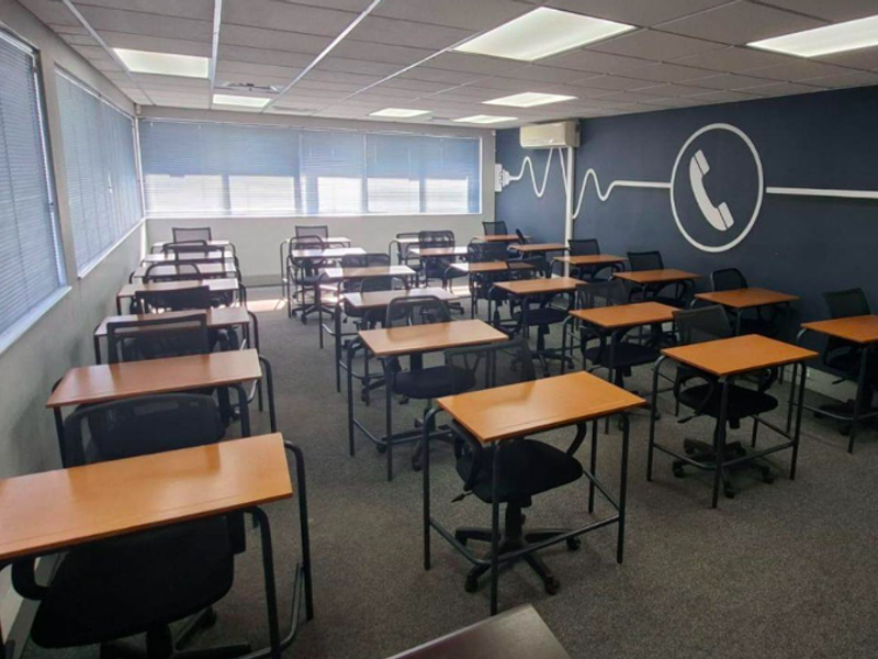 A functional office space featuring carpeted flooring, recessed ceiling lighting, wall-mounted air conditioning, and large windows with venetian blinds.