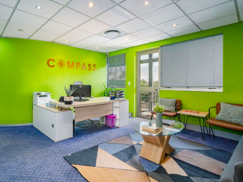 A bright office space featuring a suspended ceiling with recessed lighting, carpeted flooring, and an aluminium-framed glass door with venetian blinds.