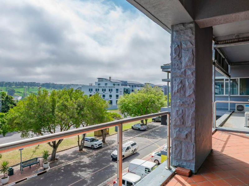 A covered balcony featuring tiled flooring, a stone-clad support pillar, and stainless steel railings, offering an elevated view of the street and surrounding area.