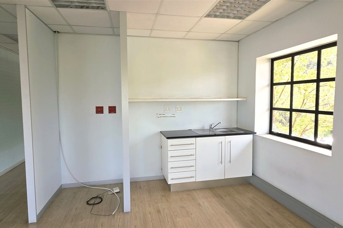 A functional office kitchenette area featuring a compact cabinet unit with a sink, laminate flooring, and a large window providing natural light.