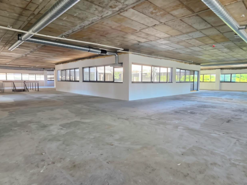 A large, open-plan office space featuring an industrial aesthetic with exposed concrete ceilings, ducting, and expansive windows providing ample natural light.
