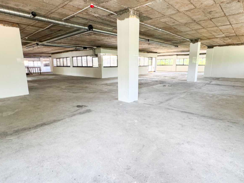 A large, open-plan office space in shell condition featuring exposed concrete ceilings, ductwork, and expansive windows providing ample natural light.