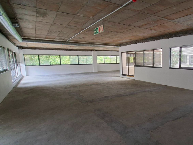Spacious open-plan office area featuring an industrial aesthetic with exposed concrete ceilings, ducting, and large windows providing ample natural light.