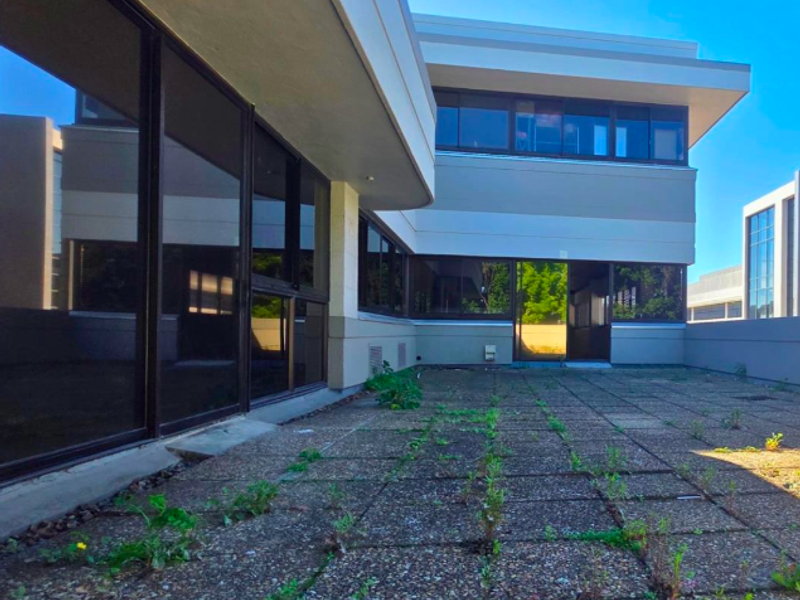 A spacious outdoor terrace area featuring paved flooring and large sliding glass doors, currently requiring maintenance to address weed growth and surface wear.