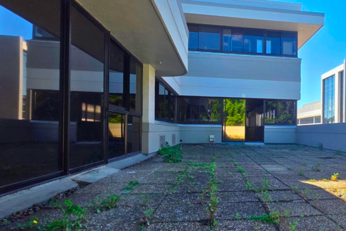 A spacious outdoor terrace area featuring paved flooring and large sliding glass doors, currently requiring maintenance to address weed growth and surface wear.