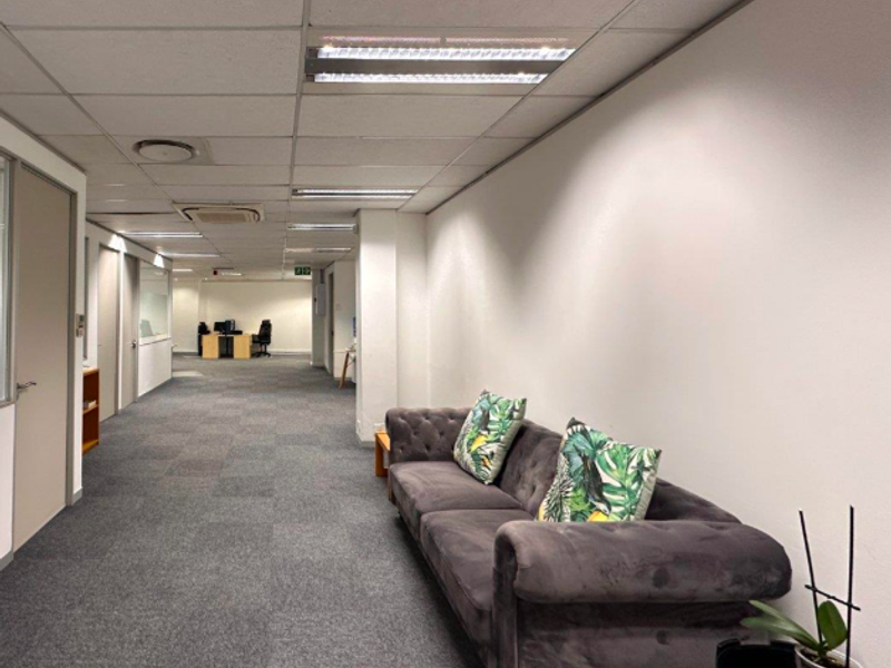 A spacious office hallway featuring a suspended ceiling with recessed lighting and neutral carpeted flooring, providing access to multiple office suites.