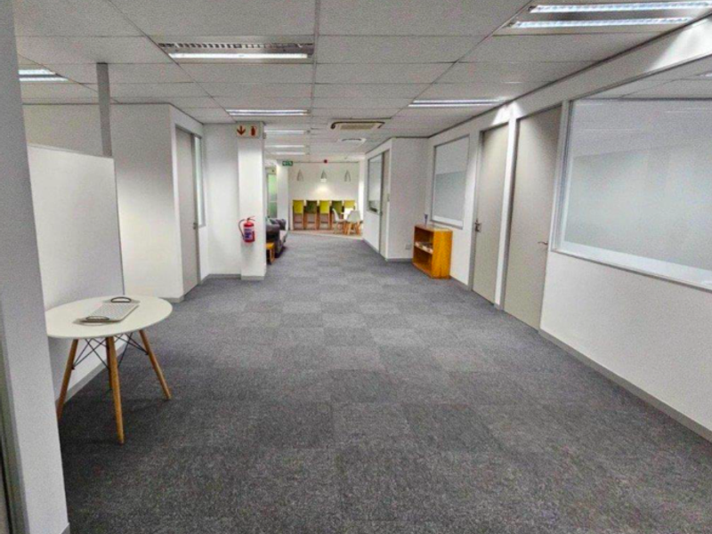 A spacious office hallway featuring carpet tiles, suspended ceilings with recessed lighting, and glass partition walls leading to private office spaces.