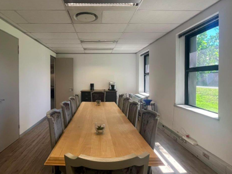 A well-lit office boardroom featuring a large wooden table, suspended ceiling with recessed lighting, and large windows providing natural light.