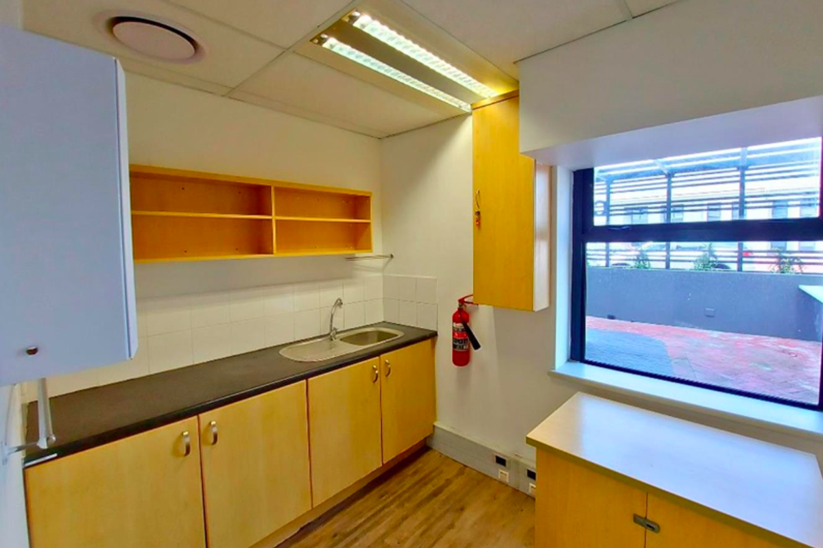 A functional office kitchenette featuring light wood cabinetry, a stainless steel sink, laminate countertops, and vinyl flooring with natural light from a large window.