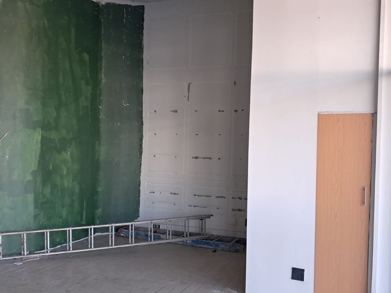 An empty industrial office space featuring large windows, a wooden door, and walls currently undergoing renovation with visible surface imperfections.