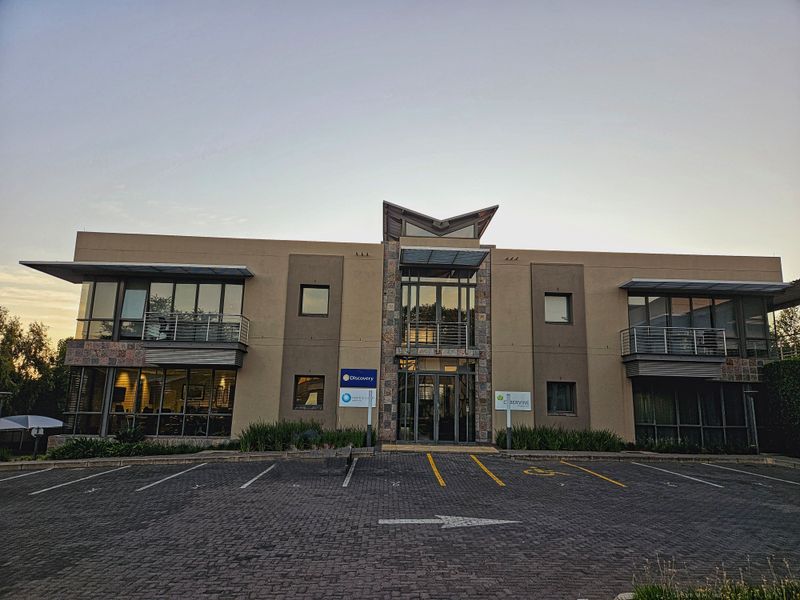 Front elevation of a modern double-storey office building featuring stone cladding, glass facade elements, metal awnings, and a paved parking area.