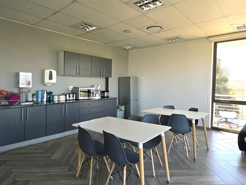 A functional office kitchenette featuring modern grey cabinetry, laminate countertops, a stainless steel sink, and large windows providing ample natural light.