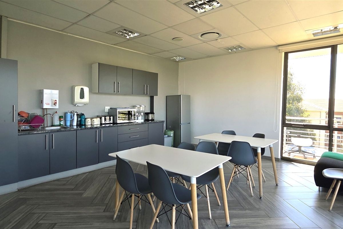 A functional office kitchenette featuring modern grey cabinetry, laminate countertops, a stainless steel sink, and large windows providing ample natural light.