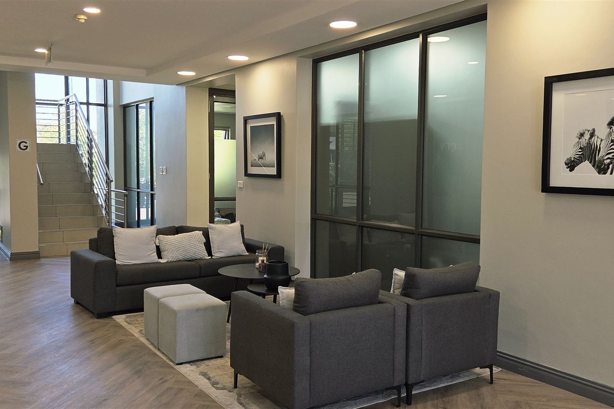 Modern office reception area featuring herringbone flooring, recessed lighting, and glass-partitioned office spaces with a staircase leading to upper levels.
