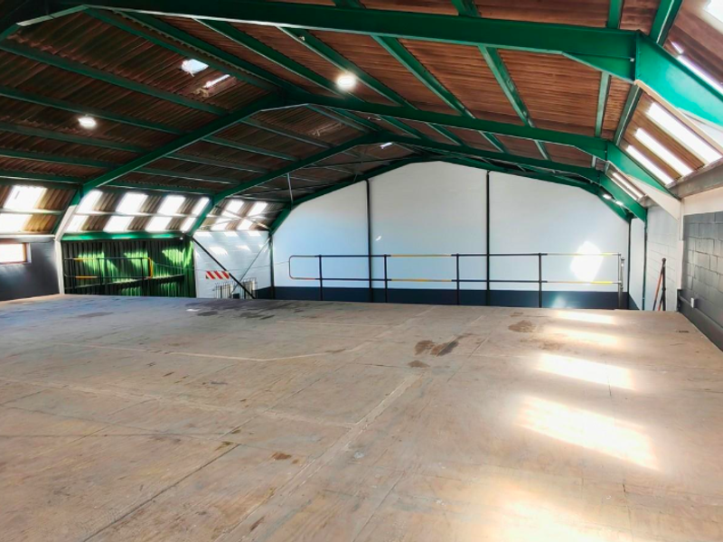 Spacious open-plan office area featuring exposed steel roof trusses, natural light from multiple skylights, and a mezzanine level with safety railing.