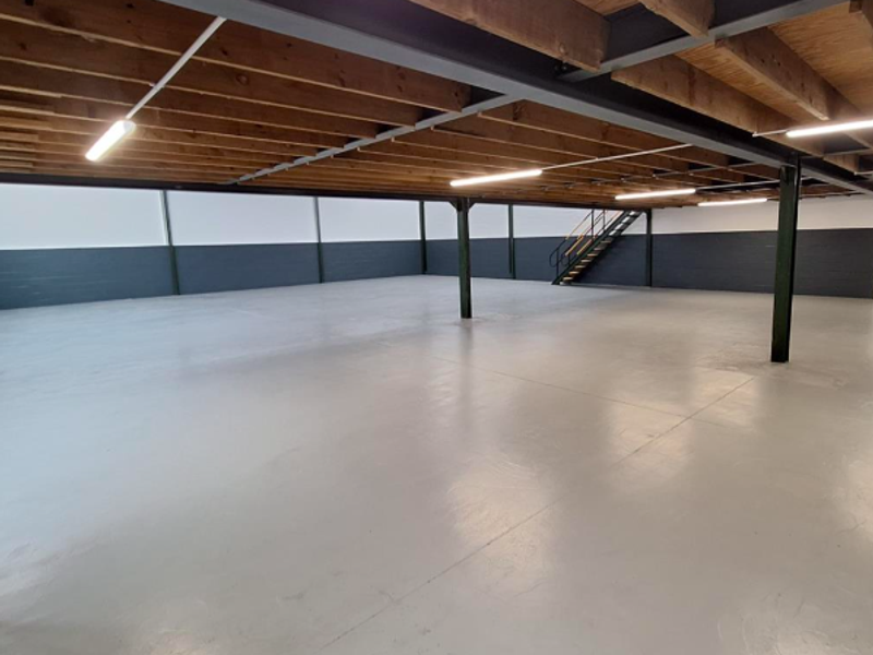 Spacious open-plan office area featuring exposed timber ceiling joists, steel structural columns, and a durable epoxy-coated concrete floor with an integrated staircase.