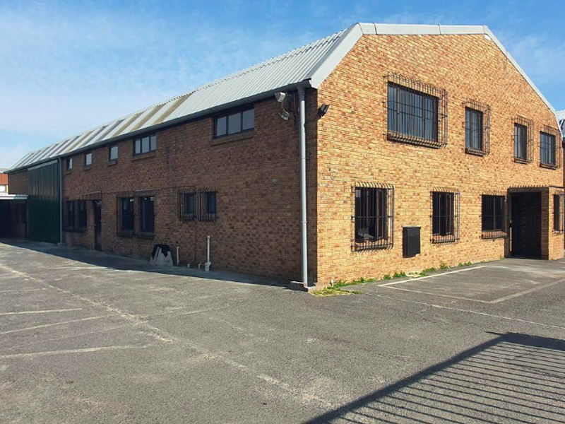 Exterior view of a multi-story brick office building featuring a metal roof, security-barred windows, and an adjacent asphalt parking area with covered bays.