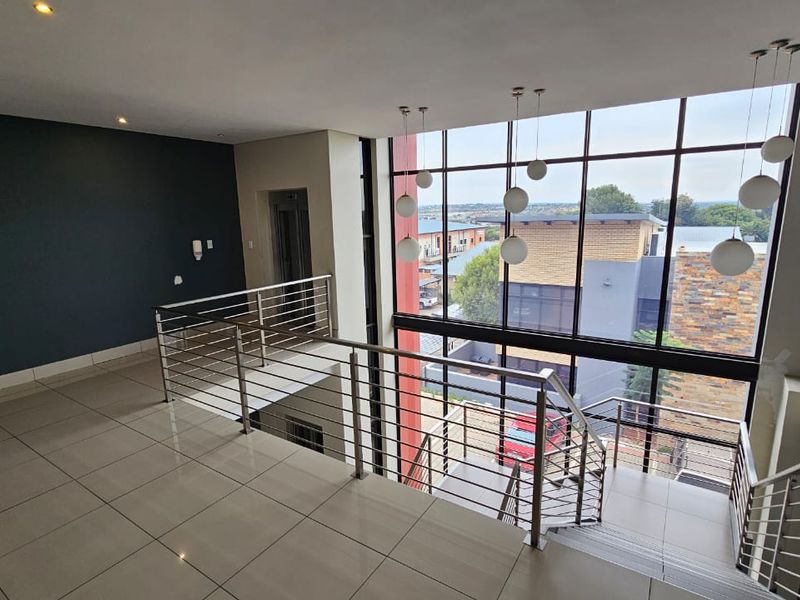 Modern office hallway featuring a double-volume glass facade, stainless steel balustrades, tiled flooring, and contemporary pendant lighting with views of the surrounding area.