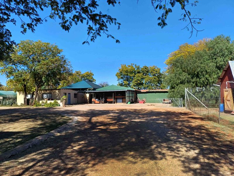 A wide view of the farm property entrance featuring a gravel driveway, covered carport area, and surrounding mature trees.
