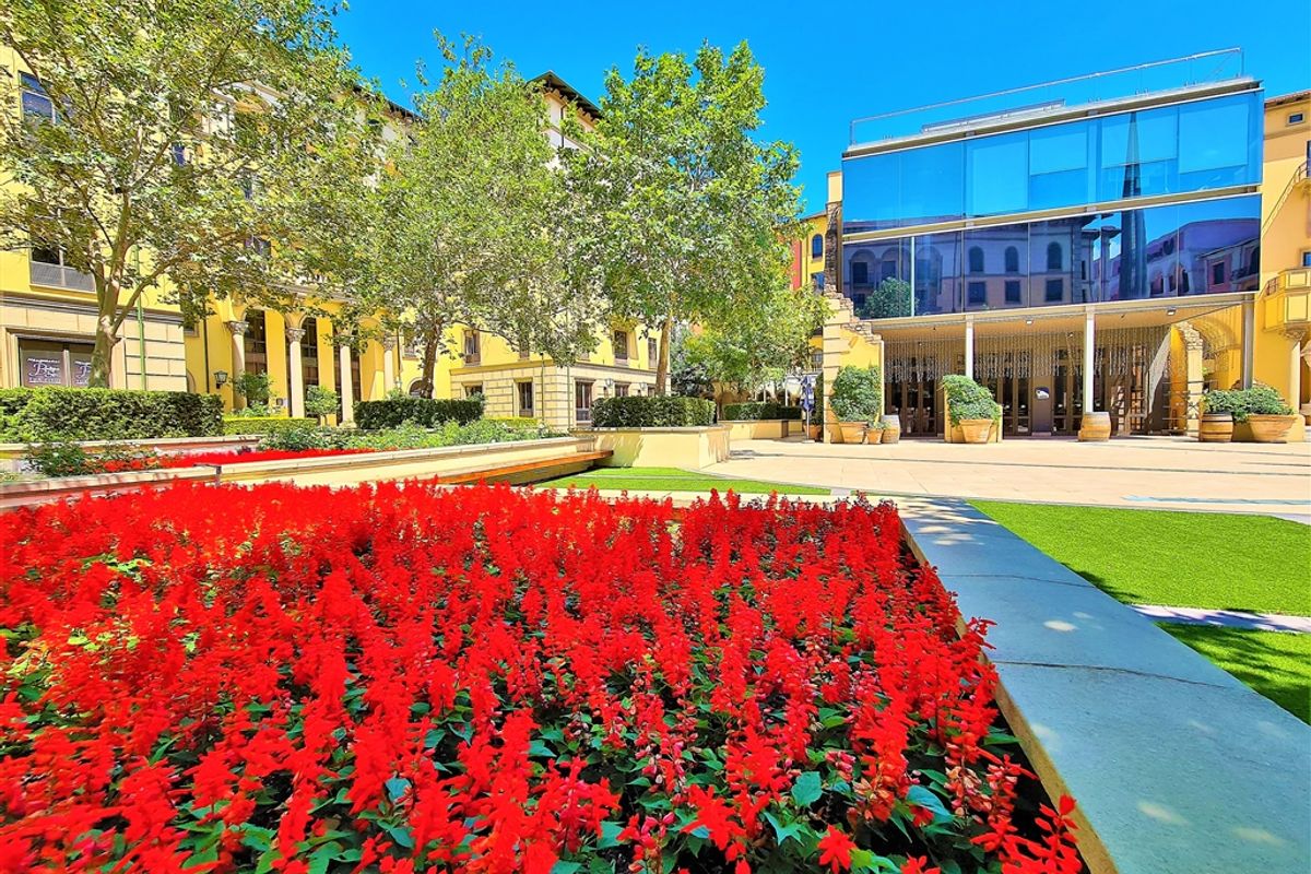 A well-maintained outdoor courtyard area featuring landscaped flower beds, manicured lawns, and modern glass-fronted office building architecture.