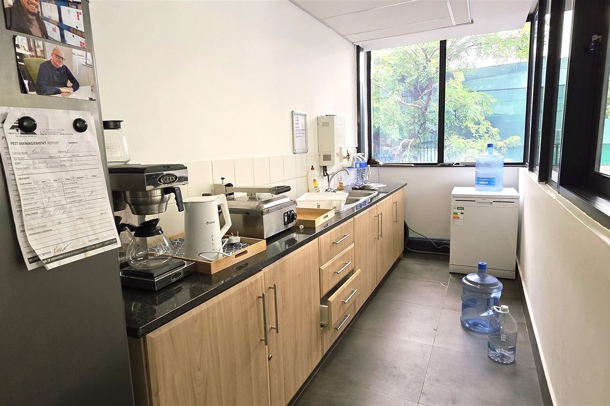 A functional office kitchenette featuring light wood cabinetry, dark granite countertops, a stainless steel sink, and large windows providing natural light.