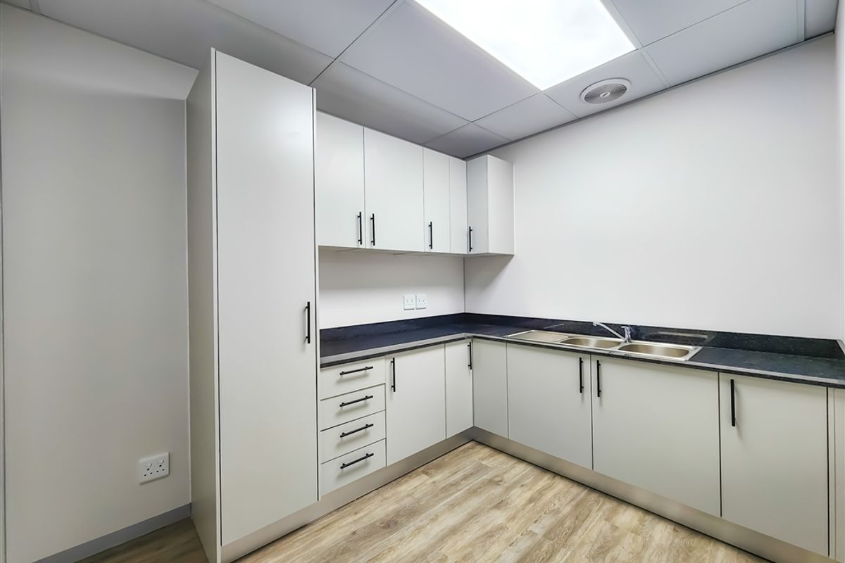 A clean, modern office kitchenette featuring white cabinetry, dark countertops, a double stainless steel sink, and wood-look flooring.