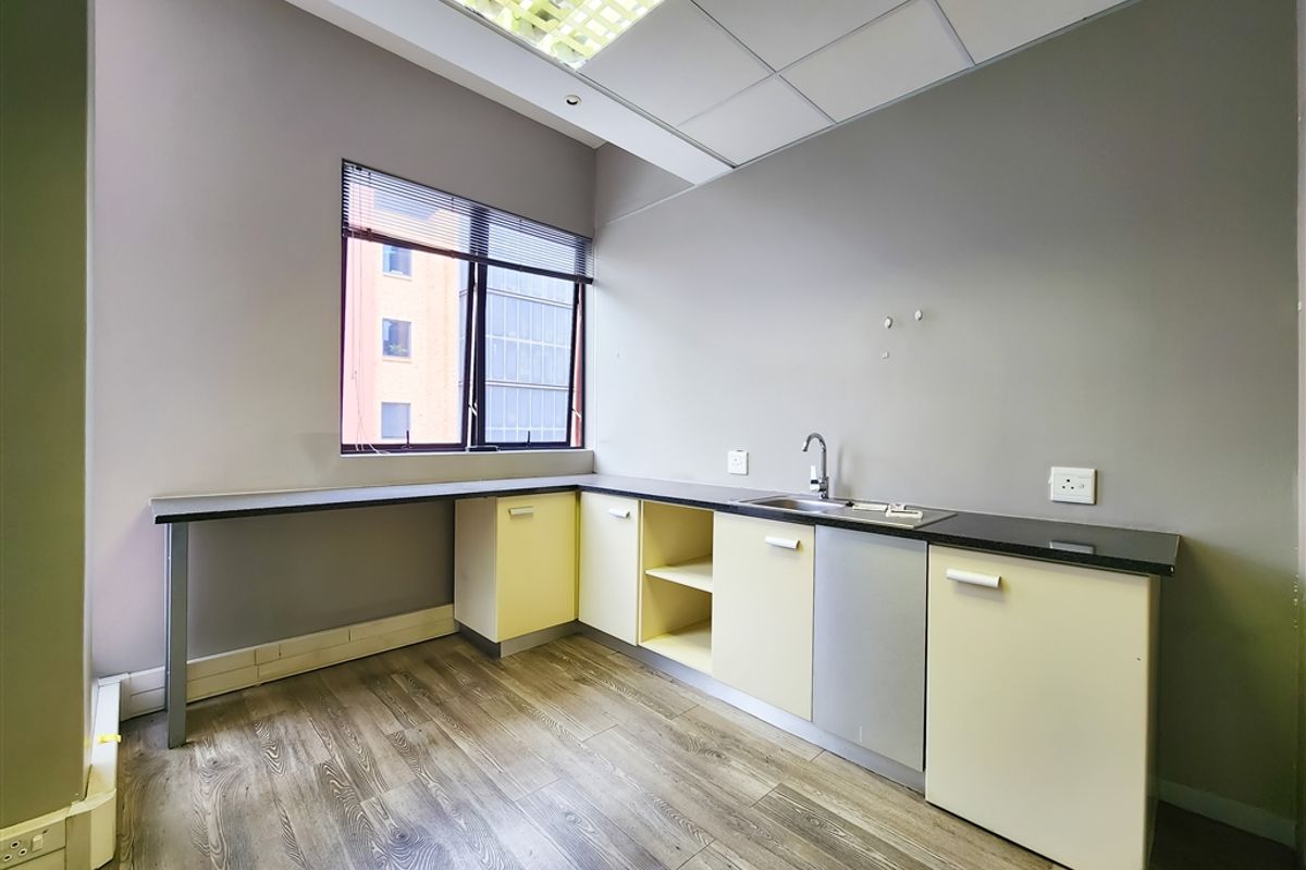A functional office kitchenette featuring built-in cabinetry, a stainless steel sink, laminate countertops, and wood-look flooring under a suspended ceiling.