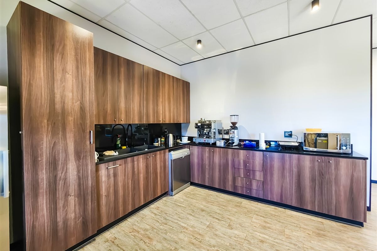 A modern office kitchenette featuring wood-grain laminate cabinetry, dark countertops, integrated appliances, and a clean, professional suspended ceiling finish.