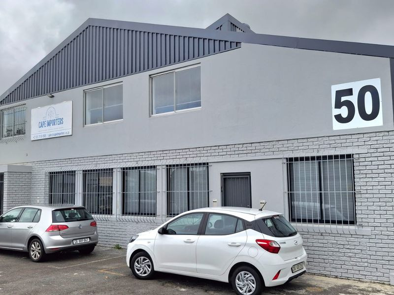 Front elevation of an industrial warehouse building featuring a grey painted facade, metal roofing, and security-barred windows with dedicated parking space.