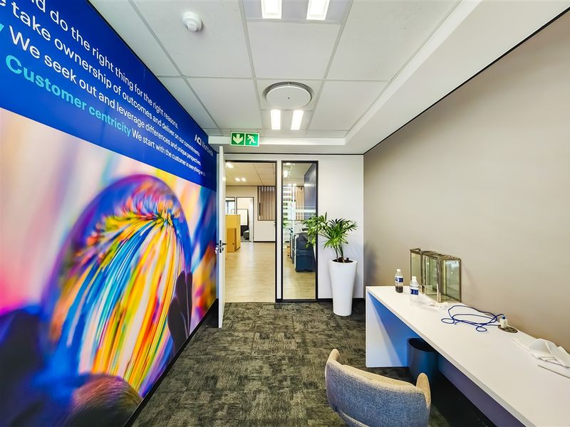 A modern office corridor featuring a vibrant branded wall graphic, recessed ceiling lighting, and a built-in desk workstation with glass partition access.