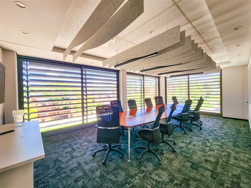 A professional office boardroom featuring acoustic ceiling baffles, large windows with external louvre blinds, and fitted carpeting.