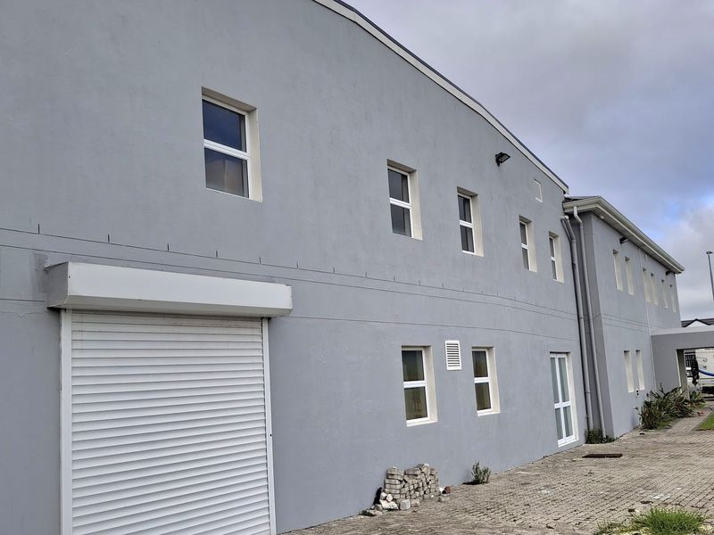 Rear exterior view of an industrial building featuring a roller shutter door, paved access area, and multiple windows along the facade.