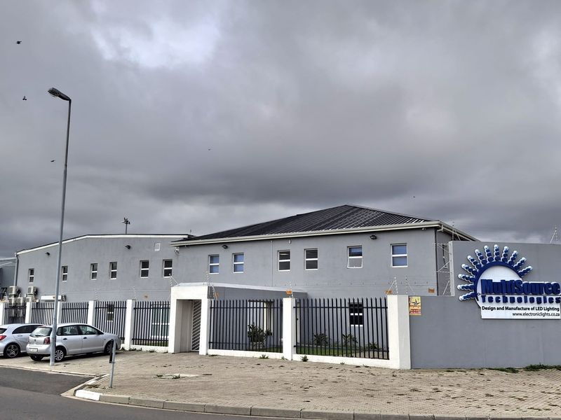 Front exterior view of an industrial facility featuring a grey facade, metal perimeter fencing with security spikes, and prominent company signage.