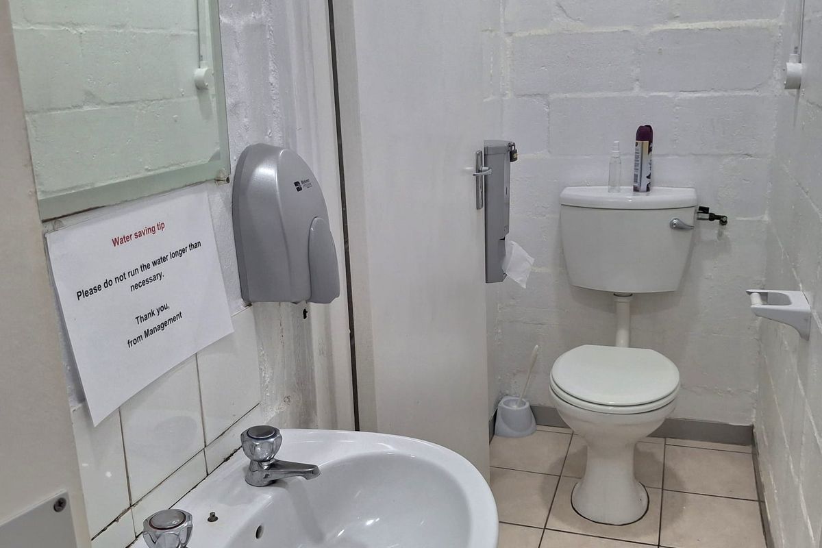 A functional industrial-style restroom featuring a pedestal basin, wall-mounted soap dispenser, and a standard toilet with tiled flooring and painted brick walls.