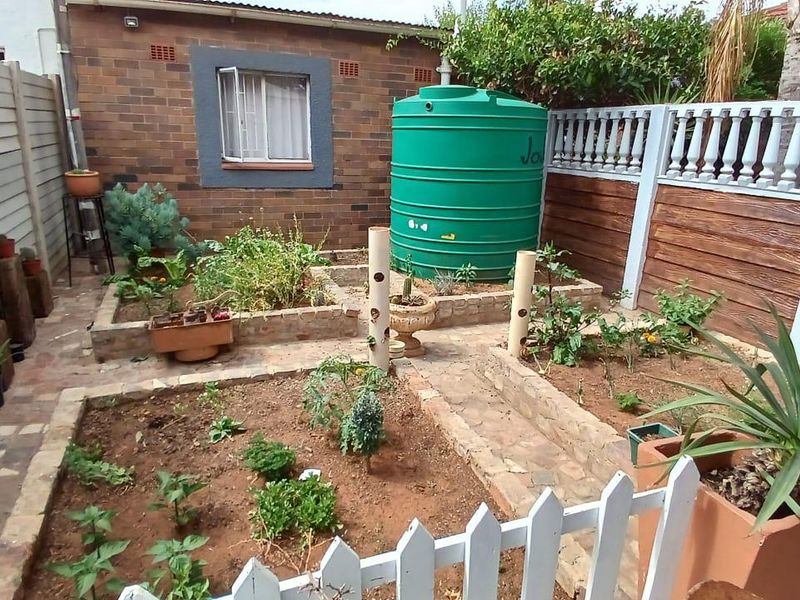 A compact rear garden featuring brick raised planting beds, paved walkways, a large green water storage tank, and secure perimeter fencing.