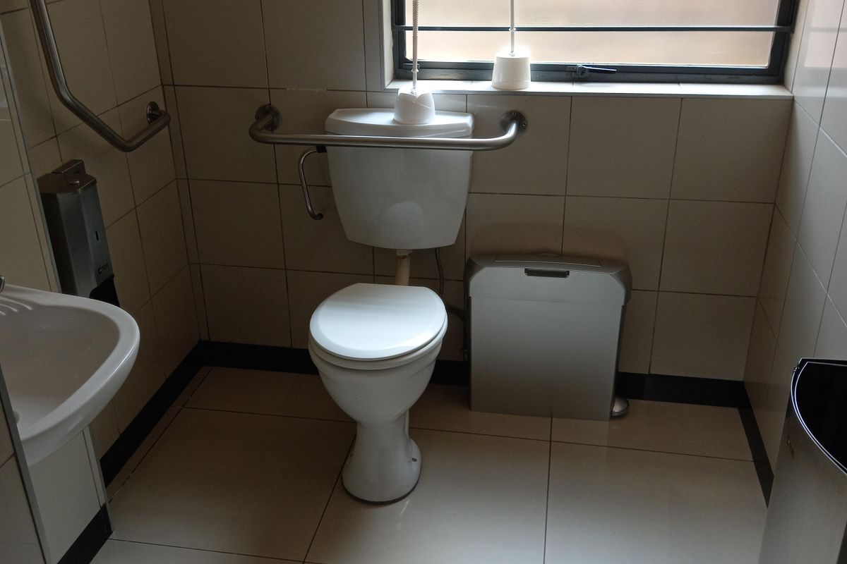 A functional office restroom featuring tiled walls and flooring, a toilet with safety grab rails, a wall-mounted basin, and a window with security bars.