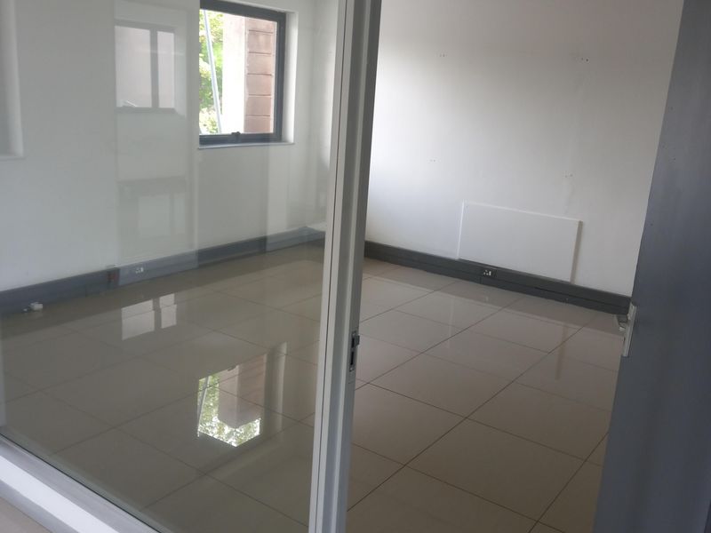 A bright, empty office space featuring tiled flooring, a glass partition wall, and a suspended ceiling with recessed lighting.
