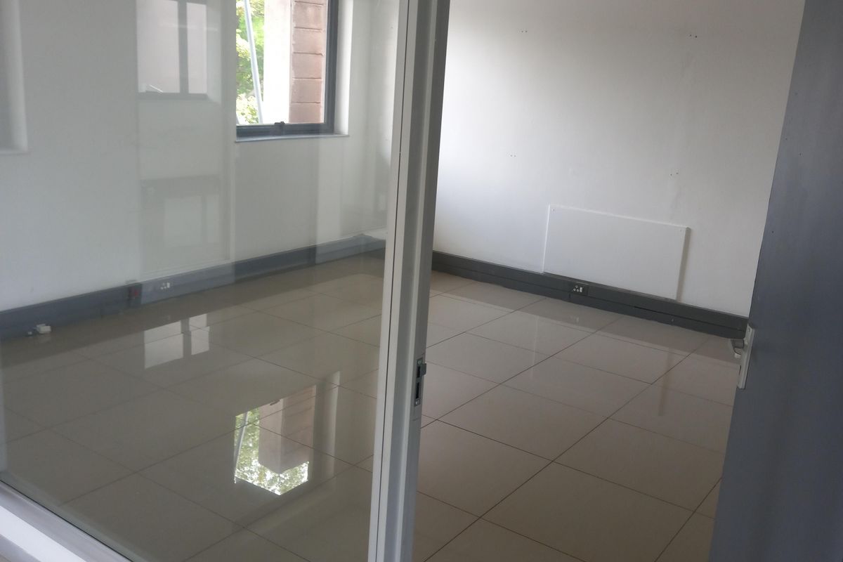 A bright, empty office space featuring tiled flooring, a glass partition wall, and a suspended ceiling with recessed lighting.
