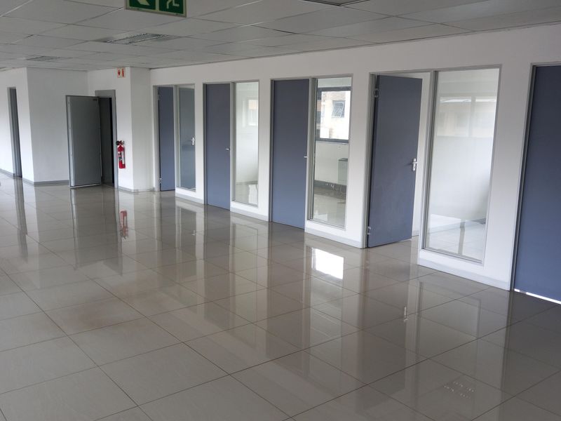 A spacious, empty office area featuring polished tiled flooring, suspended ceiling tiles, and multiple glass-paneled partition doors.